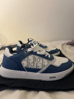 Dior sneakers blå och vit monogram - Säljer ett par Dior sneakers i blått och vitt med tydligt Dior-monogram på sidan. Skorna har snörning, perforerad tå och platt sula. Materialmix av denim och skinn ger en lyxig vibe. Grå och vit yttersula med Dior-logga undertill. Perfekt för dig som gillar exklusiva sneakers.