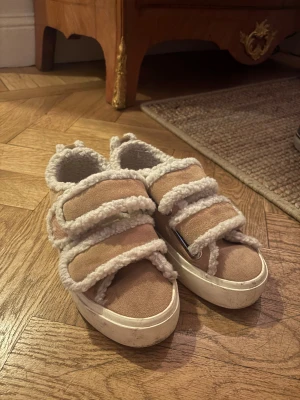 Beige sneakers från INUIKII med fårull - Unika beige sneakers från INUIKII med fluffiga detaljer i fårull och breda kardborreband. Skorna har en rund tå och vit platt sula med tydligt mönster undertill. Utsidan är i mocka och insidan är fodrad med mjuk ull för extra komfort storlek 37.