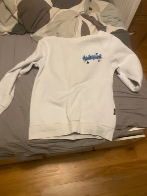 Vit hoodie från Hoodrich XS - Vit hoodie från Hoodrich i storlek XS med flera blå och svarta graffiti-inspirerade tryck på ryggen och ett mindre tryck på bröstet. Tröjan har en klassisk känguruficka framtill och är tillverkad i mjuk bomullsmix. Perfekt för dig som gillar streetwear och snygga detaljer.