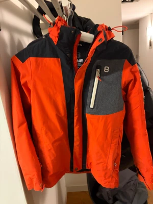 Orange och svart vindjacka från 8848 Altitude - Snygg röd och svart vindjacka från 8848 Altitude med huva och grå detalj på bröstet. Jackan har dragkedja framtill, praktiska fickor och justerbar huva. Perfekt för dig som gillar en sportig och färgstark stil.