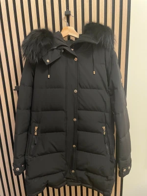 Svart dunjacka Hollies med fuskpäls - Snygg svart dunjacka från Hollies med stor huva och fluffig svart päls Jackan har guldiga detaljer, dragkedjor och knappar, samt quiltad design. Jackan köptes i slutet av vintern, använd max 4 gånger. Den är nu tyvärr för stor och kommer ej till användning så säljer den.
