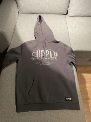 Grå hoodie från Supply & Demand - Snygg grå hoodie från Supply & Demand med stort tryck på bröstet och textdetaljer. Tröjan har huva med snörning, känguruficka och ribbade muddar. Perfekt för dig som gillar streetwear och vill ha en chill look.