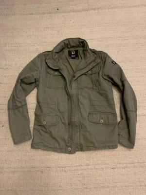 Olivgrön parkajacka från Brandit - Säljer en olivgrön parkajacka från Brandit med flera fickor framtill, både med knapp och dragkedja. Jackan har hög krage, klassisk millitärstil och är Perfekt för dig som gillar en avslappnad och stilren look.