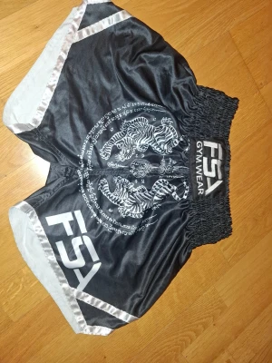 Svarta FSA träningsshorts med tigrar - Svarta träningsshorts från FSA Gym Wear i glansigt material med bred resårmidja och snörning. Framsidan har ett stort vitt tryck med två tigrar och asiatiska tecken, samt vita detaljer längs kanterna. Perfekta för gym eller kampsport.