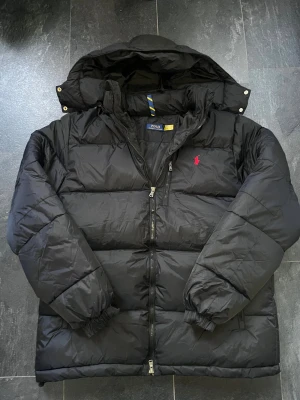 Svart pufferjacka från Polo Ralph Lauren - Svart pufferjacka från Polo Ralph Lauren med röd broderad logga på bröstet. Jackan har huva, tvåvägsdragkedja och är vadderad för extra värme. Materialet är glansig polyester och jackan har långa ärmar med resår vid handlederna.