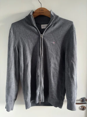 Grå kofta från GANT med dragkedja - Snygg grå kofta från GANT i storlek M, med ribbad krage och muddar. Tröjan har hel dragkedja framtill och ett broderat GANT-emblem på bröstet. Materialet är mjuk bomull och passformen är normal. Perfekt för lager-på-lager under höst och vinter.