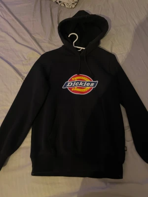 Svart hoodie från Dickies XS - Svart hoodie från Dickies i storlek XS med stort färgglatt logotryck på bröstet. Tröjan har huva med snörning och en klassisk magficka framtill. Perfekt för dig som gillar streetwear och vill ha en enkel men snygg look.