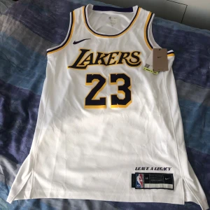 Vit Lakers baskettröja LeBron James #23 **Storlek M** - Oanvänd vit baskettröja från Nike med Lakers-logga och nummer 23 i gult och lila. På ryggen står det 'James' och 23. Tröjan är ärmlös och har klassisk rund halsringning med lila och gula detaljer i lätt och ventilerande material.