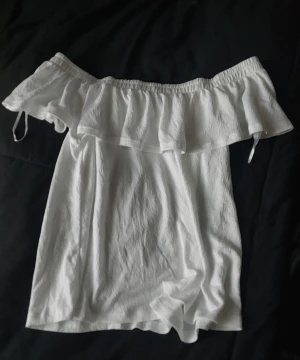 Vit offshoulder tröja från Gina Tricot - Superfin vit tröja  från Gina Tricot med offshoulder-modell och volang upptill. Tröjan är i ett lätt, krinklat bomullstyg och har en avslappnad passform. Perfekt för varma dagar när du vill känna dig både söt och bekväm.