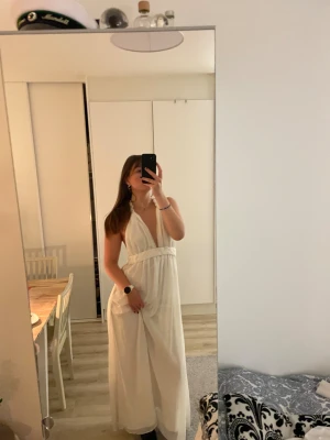 Vit långklänning med flätad midja - Säljer en vit långklänning med djup v-ringning och flätad detalj i midjan. Klänningen har axelbandslös design med knyt i nacken och är tillverkad i ett lätt, skirt material som ger ett flowy fall. Dragkedja i sidan för enkel påklädning. Finns ingen lapp med storlek eller märke men passar mig, M.