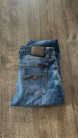 Blå jeans från Nudie Jeans - Säljer ett par klassiska blå jeans från Nudie Jeans. Modellen har och fem fickor, med Nudie Jeans logga på bakfickan. Jeansen är i stretchigt denim och har knappgylf.