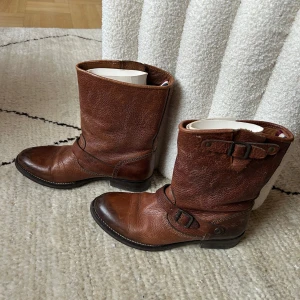 Bruna skinnboots från Hilfiger Denim - Snygga bruna boots i äkta skinn från Hilfiger Denim. Modellen har rund tå, platt sula och coola spänndetaljer både upptill och vid ankeln. Perfekta till jeans eller kjol för en avslappnad men stilren look.