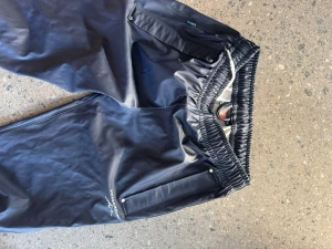 Svarta regnbyxor från Swedmount - Svarta regnbyxor från Swedmount med elastisk midja och snörning. Byxorna har raka ben, två fickor med dragkedja och tryckknappar vid benslut. Materialet är syntet och vattenavvisande, perfekt för utomhusaktiviteter.