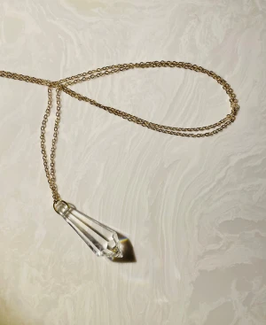 Guldhalsband med kristallpendant - Unikt halsband med tunn guldfärgad kedja och en stor, facetterad kristallpendant i transparent glas. Kristallen har en cool geometrisk form som fångar ljuset och ger snygga reflektioner. Perfekt statement-piece för dig som gillar stilrena och moderna smycken.