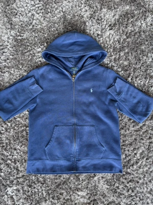 Blå hoodie från Polo Ralph Lauren - Snygg blå hoodie från Polo Ralph Lauren med dragkedja och huva. Tröjan har korta ärmar, fickor framtill och den klassiska Polo-loggan broderad på bröstet. Perfekt för chill dagar och enkel att matcha med jeans eller joggers.