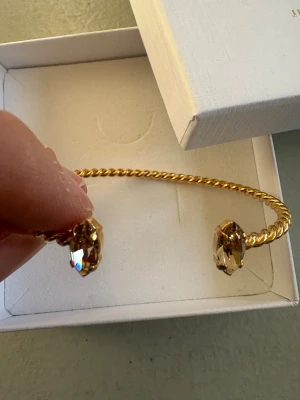 Guldarmband med kristaller Caroline Svedbom - Snyggt armband från Caroline Svedbom i guldtonad metall med tvinnad design. Armbandet är öppet och pryds av två gnistrande, ljusa kristaller i båda ändar som ger en lyxig känsla. Perfekt accessoar för att lyfta din outfit.