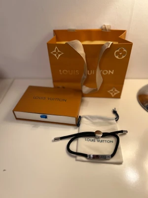 Louis Vuitton armband - Stilrent svart armband från Louis Vuitton med silverfärgade detaljer och logotyp på metallbrickan. Armbandet har justerbar passform med rund låsdetalj och levereras i originalpåse och ask. Perfekt accessoar för dig som gillar lyxiga och minimalistiska smycken.