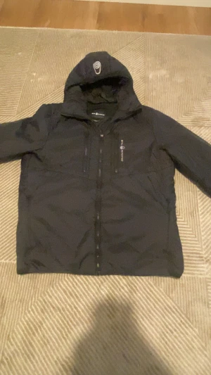 Svart pufferjacka från Sail Racing - Svart pufferjacka från Sail Racing med huva och flera dragkedjor framtill. Jackan har en sportig look, logga på bröstet och är tillverkad i glansig polyester. Perfekt för kalla dagar och har en modern, clean stil.