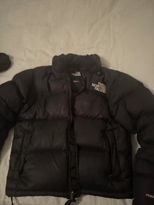 Svart dunjacka från The North Face - Säljer en svart pufferjacka från The North Face med klassisk logga på bröstet. Jackan är vadderad med dun, har hög krage och dragkedja framtill. Perfekt för kalla dagar och riktigt snygg streetstil. Äkta