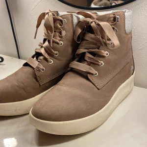 Beige boots från Timberland strl 37 - Snygga beige boots från Timberland med silvrig vadderad kant och breda satinliknande snören. Skorna har en vit platt sula och är tillverkade i skinn. Perfekta för dig som vill ha en stilren och bekväm look.