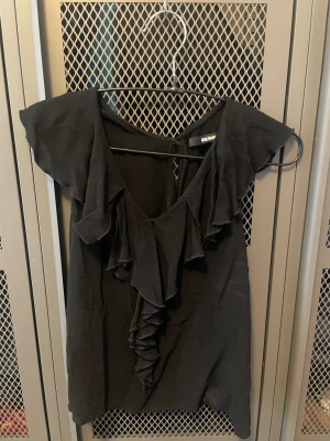 Svart volangblus från Bik Bok XS - Svart blus från Bik Bok i storlek XS med djup v-ringning och volangdetaljer framtill. Ärmlös modell med knytning i nacken och lätt, flowigt tyg som ger en snygg siluett. Perfekt för dig som gillar trendiga och feminina detaljer.