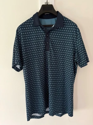 Mönstrad blå kortärmad golfpiké från Galvin Green - Snygg blå pikétröja från Galvin Green med vitt geometriskt mönster över hela plagget. Klassisk krage, knappar framtill och korta ärmar. Perfekt för dig som gillar sportig och stilren look. Storlek: L passar M. Nypris: 999kr