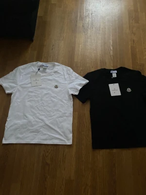 Moncler t-shirt svart & vit M - Två stilrena Moncler t-shirts i svart och vit med klassisk rund hals och korta ärmar. Båda har Moncler-loggan broderad på bröstet och är gjorda i mjuk bomull. Perfekt för dig som gillar exklusiva och enkla plagg med snygg branding.