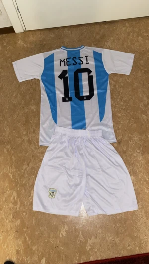 Argentina Messi #10 fotbollströja set - Säljer ett Argentina fotbollströja och shorts set med Messi #10 på ryggen. Tröjan är vit med ljusblå vertikala ränder, kortärmad och har AFA-emblem på bröstet. Materialet är lätt och andas, perfekt för match eller träning. Klassisk rund hals och snygga detaljer på axlarna.