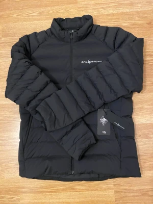 Svart dunjacka från Sail Racing - Svart dunjacka från Sail Racing med stilren design och diskret logga på bröstet. Jackan har horisontella sömmar, hög krage och dragkedja framtill. Perfekt för kalla dagar och enkel att matcha med allt. Snygg och funktionell jacka för dig som gillar sportig stil. Den är helt ny med lappar kvar jag köpte fel storlek och kunde inte lämna tillbaka den