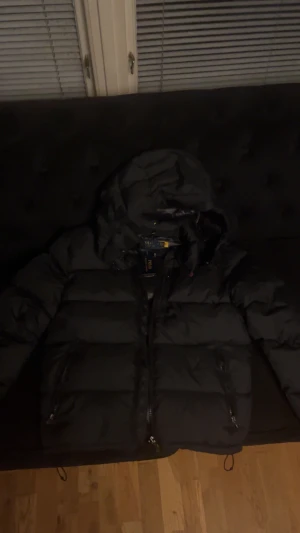 Svart dunjacka från Polo Ralph Lauren - Säljer en svart dunjacka från Polo Ralph Lauren med huva och dragkedja framtill. Jackan har två sidofickor med dragkedja och en klassisk pufferdesign som håller dig varm under kalla dagar. Perfekt för dig som vill ha en stilren och funktionell vinterjacka.