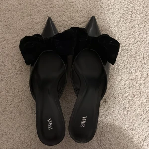 Svarta pumps med rosett från Zara - Snygga svarta pumps från Zara i super skick med spetsig tå och låg klack. Skorna har en glansig finish och är dekorerade med en stor svart rosett i sammet framtill. 