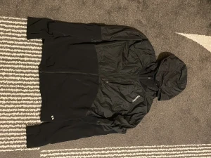 Svart vindjacka från Röhnisch med huva - Svart vindjacka från Röhnisch med hel dragkedja och huva. Jackan har en sportig look med paneler i blankt och matt material, samt diskret logga på bröstet. Perfekt för blåsiga dagar och enkel att matcha med allt.