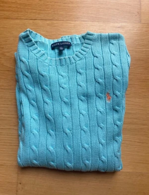 Ljusblå kabelstickad tröja Ralph Lauren - Snygg ljusblå kabelstickad tröja från Ralph Lauren med rund hals och lång ärm. Tröjan har det klassiska broderade Polo-logot på bröstet och ribbade muddar vid ärmslut och nederkant. Perfekt för dig som gillar preppy stil och vill ha något som sticker ut.