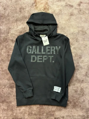 Svart hoodie från Gallery Dept. - Svart hoodie från Gallery Dept. med stor logotyp på bröstet och en vit patch med text på ärmen. Tröjan har huva, ribbade muddar och en klassisk känguruficka framtill. Materialet är mjuk bomull, perfekt för chill dagar och streetwear vibes.