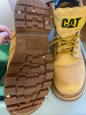 CAT boots i beige skinn, storlek 38 - Säljer ett par klassiska CAT boots i beige skinn med grov sula och rund tå. De har gul- och brunmönstrade snören, vadderad svart krage och tydlig CAT-logga på plösen. Perfekta för dig som gillar robust stil och vill ha slitstarka skor. Eftersom de bara stått i garderoben så har skinnet i kragen på högra skon blivit torrt. Jag har efteråt smörjt in den med skokräm.
