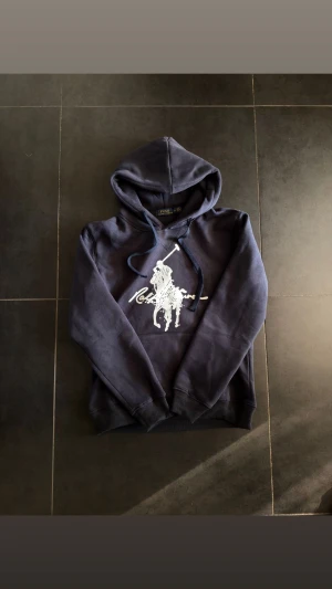 Mörkblå hoodie från Polo Ralph Lauren - Snygg mörkblå hoodie från Polo Ralph Lauren med stor vit logga på bröstet. Tröjan har huva med snörning och ribbade muddar vid ärmslut och nederkant. Tillverkad i mjuk bomull, perfekt för chill dagar och streetstyle.