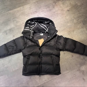 Burberry Puffer - Svart dunjacka från Burberry med klassisk Säljer en elegant och mångsidig Burberry-jacka i svart, i nyskick. Dunfodrad för värme och komfort med det klassiska rutmönstret i huvan som ger en exklusiv touch. Praktiska dragkedjefickor, avtagbar huva och diskret logopatch på ärmen. Passar S/M. Jackan är i princip som ny och utan defekter. En lyxig kombination av stil och funktion – perfekt för både vardag och tillfällen då du vill klä upp dig.  📦Snabb leverans - skickas samma dag📌.