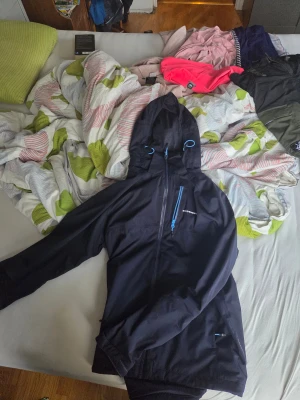 Svart vindjacka Swedemount L - Svart vindjacka från Swedemount i storlek L. Jackan har huva med blå detaljer, hel dragkedja framtill och en bröstficka med blå dragkedja. Materialet är syntet och jackan är perfekt för blåsiga dagar. Snygg och funktionell design med Swedemount-logga på bröstet.