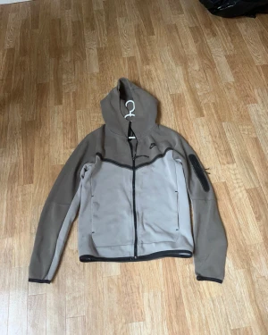 Beige hoodiejacka från Nike XS - Nike hoodiejacka i beige och bruna toner med svart dragkedja och detaljer. Jackan har huva, lång ärm och en ficka med dragkedja på ena ärmen. Tillverkad i mjukt syntetmaterial, perfekt för chill eller streetwear. Svårt att hitta denna färgen!