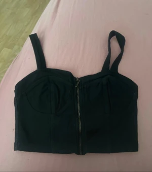 Svart korsett-topp med dragkedja - Svart korsett-topp med breda axelband och snygg dragkedja framtill. Toppen har markerade kupor och är croppad i modellen. Perfekt för att ge din outfit en edgy vibe. Materialet känns som bomull med lite stretch för skön passform.