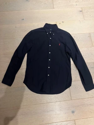 Svart skjorta från Ralph Lauren M - Svart slim fit skjorta från Ralph Lauren med klassisk krage och vita knappar. På bröstet finns den ikoniska röda Polo-loggan broderad. Skjortan är långärmad och tillverkad i mjuk bomull, perfekt för en clean och stilren look.