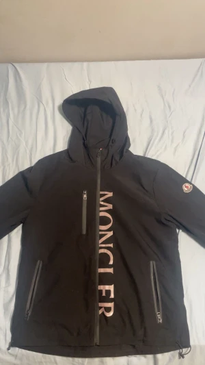 Svart vindjacka från Moncler - Svart vindjacka från Moncler med stor logga framtill och emblem på ärmen. Jackan har huva, tre dragkedjefickor och snygga detaljer med Monclers klassiska färger på insidan. Perfekt för dig som gillar stilren och sportig look.