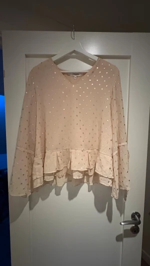 Ljusrosa blus med volang och prickar - Supersöt ljusrosa blus från Kappahl med glittriga guldprickar, volangkant nedtill och långa ärmar med knyt vid handlederna. Blusen har en luftig passform och är gjord i ett lätt, transparent material som ger en romantisk vibe.