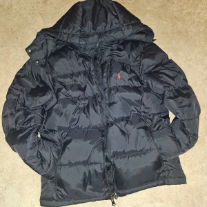 Svart pufferjacka från Polo Ralph Lauren - Svart pufferjacka från Polo Ralph Lauren med avtagbar huva och klassisk röd logga på bröstet. Inte använt så mycket, passar inte riktigt min stil. Fick den som present på födelsedag.