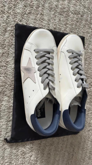 Golden Goose Superstar sneakers - Säljer ett par Golden Goose Superstar sneakers i vitt skinn med grå mockastjärna på sidan, grå snörning och mörkblå häl. Snygg vintage-inspirerad look med platt sula och rund tå. Perfekt för dig som gillar italiensk design och streetstyle.
