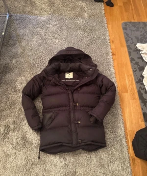 Svart dunjacka från Boomerang S - Säljer en svart dunjacka från Boomerang i storlek S. Jackan har huva, dragkedja och knappar framtill samt två fickor på sidorna. Klassisk pufferstil med quiltade sektioner och justerbar nederkant. Perfekt för kalla dagar. Och den har vissa hål men det syns knappt 