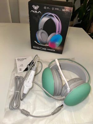 AULA Mountain S505 gaming hörlurar  - AULA Mountain S505 är ett trådbundet gamingheadset med USB-anslutning, justerbar mikrofon och bekväma over-ear-kuddar. Snygg design med färgade kåpor och RGB-belysning. Headsetet är i nyskick och levereras komplett med originalkartong och manual.AULA S505 gaming hörlurar med mikrofon. 🎁 