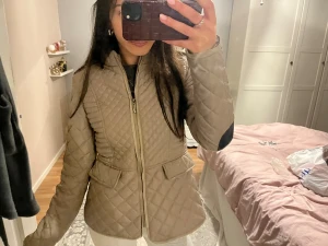 Beige quiltad jacka med svarta detaljer - Säljer en beige quiltad jacka med snygga svarta detaljer på armbågarna och dragkedja framtill. Jackan har figurnära passform, fickor med lock och markerad midja. Perfekt för dig som gillar stilren och chill look under höst och vår.