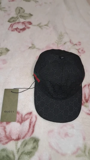 Svart Gucci keps med GG-mönster - Snygg svart keps från Gucci med klassiskt GG-monogram över hela kepsen och ikoniska gröna och röda ränder på sidan. Justerbar rem baktill för perfekt passform. Tillverkad i Italien och gjord av bomull. Perfekt för dig som vill ha en lyxig streetstyle-look. Helt ny oanvänd me etikettet.