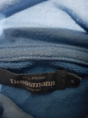 Blå hoodie från Dressmann med huva - Blå hoodie från Dressmann i storlek S. Tröjan har en klassisk huva med snörning och en stor magficka framtill. Mjuk och skön bomullskänsla, perfekt för chill dagar. Långärmad och enkel design utan mönster eller tryck.